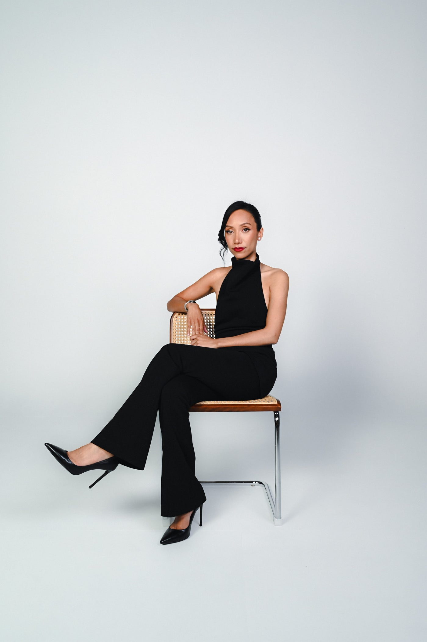 A woman wearing a black halter-neck jumpsuit and black pointed-toe heels sits on a modern wooden chair with a metal frame against a light gray studio background.